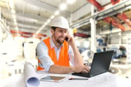 Industriearbeiter mit Helm und Warnweste vor Notebook im Einsatz in einer Industrie Halle