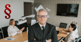 Ein älterer Herr im Anzug versinnbildlicht hier den CEO zum bildlichen Unterstreichen des Titels Geschäftsführerhaftung, im Hintergrund sitzen Mitarbeiter